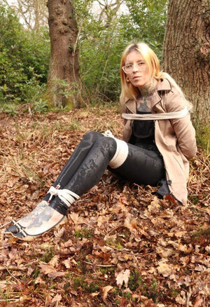 Babe with glasses can't stand up because she is hogtied by the tree